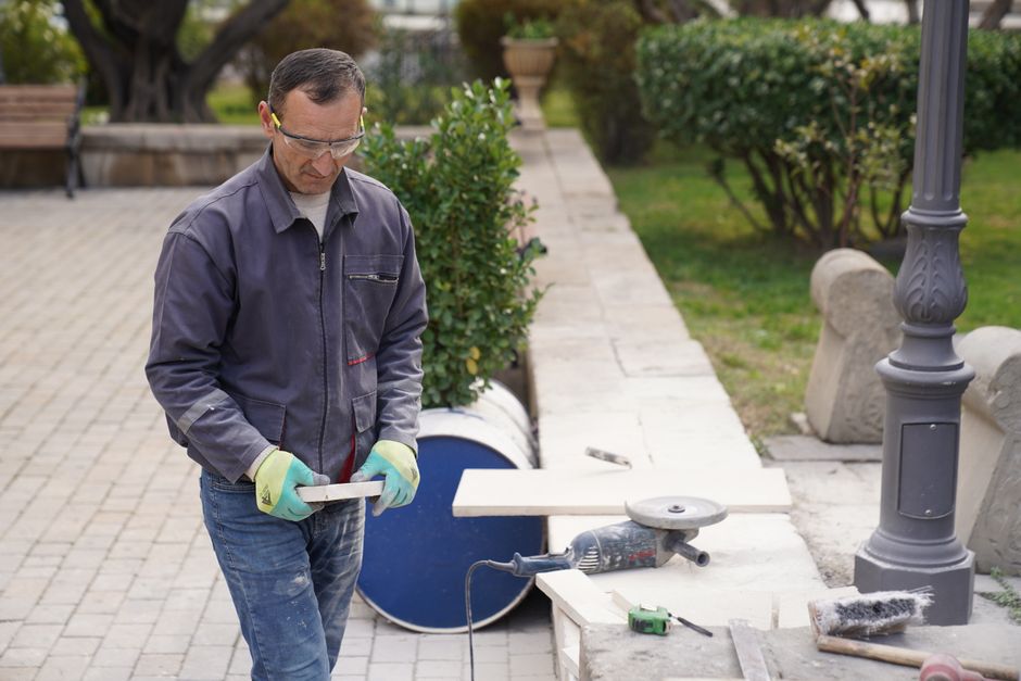 A carpenter wearing safety gear works with tools on an outdoor project in a park setting.